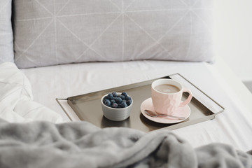 Coffee with milk in a pink cup and blueberry on a metal tray served in a bed. Light morning breakfast. Weekend relax