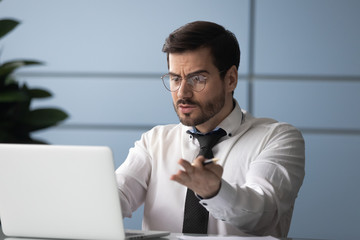 Young angry businessman outraged by news using laptop. Serious employee stressful due to report by...