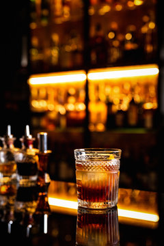 A cocktail in an old fashioned glass on a bar counter with a reflection, bottles with bitters, bokeh lights, vertical photo