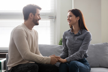Happy young man holding attractive woman hands on couch. Smiling romantic couple enjoy time...
