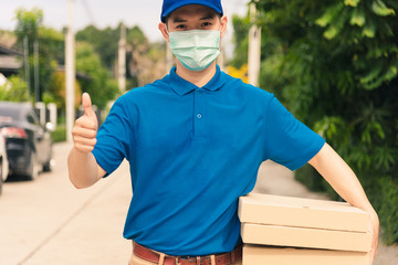 Asian young delivery man courier sending and holding fast food pizza boxed in uniform he protective face mask show thumb up for good sign, under curfew quarantine pandemic coronavirus COVID-19