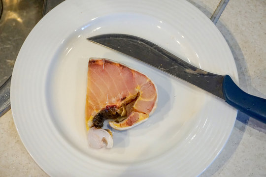 Cut Piece Of Fish, Sturgeon, Kaluga, Gourmet Meat. A Piece With Pieces Of Fat And Blood, In The Skin, Lies On A Light White Plate With Knife, Top View, Flat Lay 