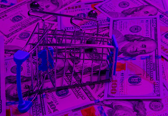 Shopaholic concept. Shopping cart on the background of one hundred dollar bills in pink neon light