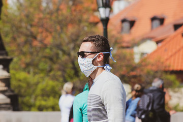 Young man with sunglasses and sewed fabric face mask photographed on the Charles Bridge in Prague, Czech Republic. Blurred people in the background. Travelling, tourism during coronavirus. COVID-19