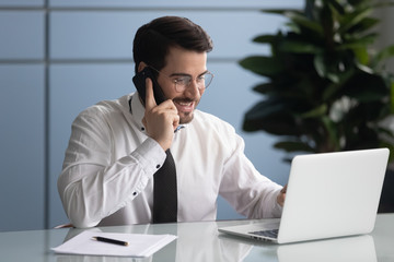 Happy businessman talking on smartphone looking at laptop screen. Smiling successful man manager make online success deal using modern computer. Employee at work.