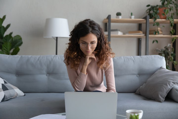 Front view pleasant smiling young woman in glasses sitting on sofa, looking at laptop screen....