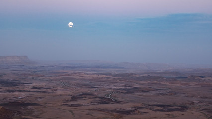 full moon peaking through clouds above the makhtesh ramon crater in israel just after sunset in...
