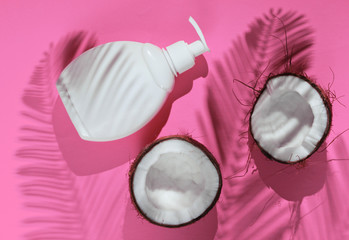 Minimalistic beauty still life. Two halves of chopped coconut and white bottle of cream with shadows from palm leaves on pink background. Creative fashion concept. Top view.