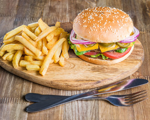 Fresh delicious Burger, bacon, cheese, tomato, French fries, onion and salad on a wooden table with a knife and fork