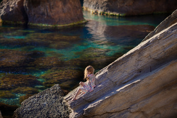 A little pretty girl in a dress stands on a large stone by the sea. A child on a background of turquoise water and rocks, a summer walk on the background of a beautiful landscape