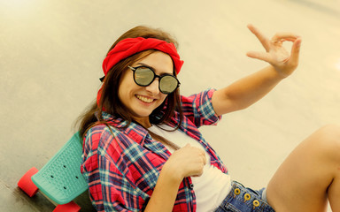 Young hipster girl in stylish mirrored glasses, shorts, red plaid shirt and bandage on her head lies with skateboard on concrete surface in skatepark