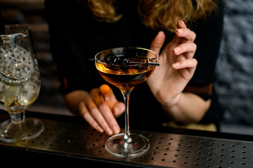 glass with cocktail which decorated small sword with cherries standing on bar
