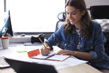 Happy young woman writing something down while working in the office. © ty