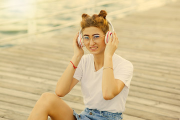 Young woman dressed in stylish clothes sits on the beach promenade and listen music in headphones