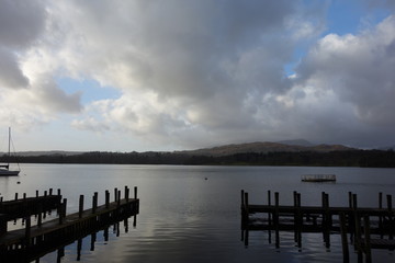 Lake with a jetty in the foreground and a hill in the background and clouds in the blue sky