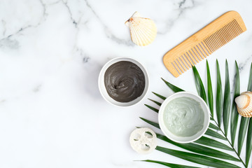 Green and black face clay masks with tropical palm leaf, hair comb, loofah sponge and seashells on marble background. SPA natural organic cosmetics for face or hair treatment, skin care