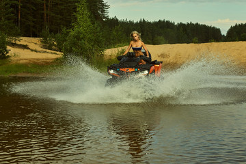 Beautiful blonde girl on a ATV rides on water and sand mountains. Green forest all around. In summer season