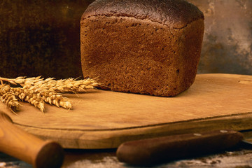 Decoration made of loaf of rye bread rye spikelets wooden board rolling pin knife with wooden holder in vintage rustic style. Old brown surface background. Closeup front view horizontal layout