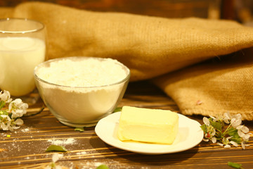 Bowl with flour, a glass of milk, eggs, butter, homemade cakes and flowers on an old wooden background. Ingredients for cooking. Black and white. The near plan. Healthy eating concept