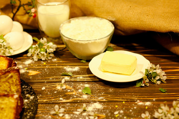 butter, a bowl with flour, a glass of milk, eggs, homemade cakes and flowers on an old wooden background. Ingredients for cooking. Black and white. The near plan. Healthy eating concept