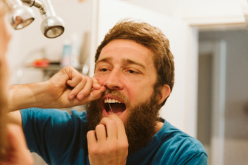 Handsome bearded man looking at mirror while using dental floss in bathroom 