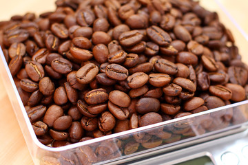 Beautiful dark aromatic freshly roasted coffee beans are poured into transparent plastic food container for weighing on silver electronic digital scale, against light brown wooden surface