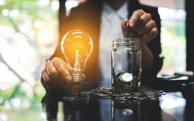 Businesswoman holding a light bulb over coins stack on the table while putting coin into a glass...