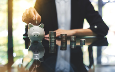 A businesswoman putting coin into piggy bank with coins stack on the table for saving money and...