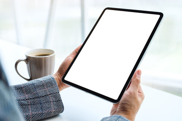 Mockup image of hands holding black tablet pc with blank white screen with coffee cup on the table