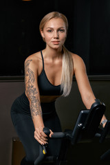 Fototapeta na wymiar Young confident woman athlete exercising on bicycle indoors. Attractive determined fitness girl doing cycling exercises in dark gym. Functional workout training of sports girl. Cardio training