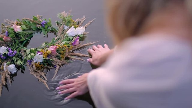 close-up of a girl drops a wreath of flowers and herbs into the water.  hands of woman near river or lake on Ivan Kupala.  bouquet of wildflowers and dried grass. female back and blond long hair