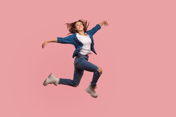 Full length of delighted happy stylish girl in checkered shirt and ripped jeans dancing in air, celebrating success, feeling free and inspired in flight. indoor studio shot isolated on pink background