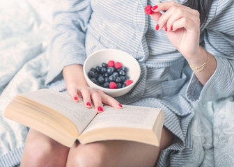 Young dark haired woman holding and reading her loved book in the white bed wearing bathrobe and eating fresh fruit salad, red nails