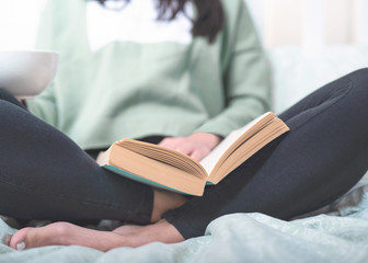 Young dark haired woman is sitting in the bed and reading her favorite book, drinking coffe from white cup