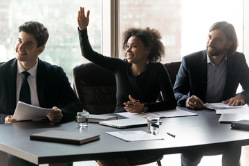 Smiling African American businesswoman raising hand, asking question to coach mentor at briefing, sitting at table in boardroom, employee engaged in team building activity at corporate meeting