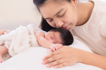 Fototapeta na wymiar Selective Focus, Asian mother kiss adorable newborn while sleep, mom support baby girl with love, care, protection hold hand together. Motherhood lifestyle take care infant on bed at home concept