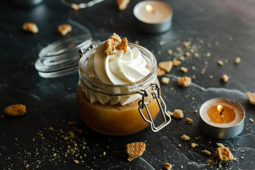 Delicious dessert in a jar. Banana dessert with caramel and whipped cream, garnished with cookies and nuts. Romantic composition with candles
