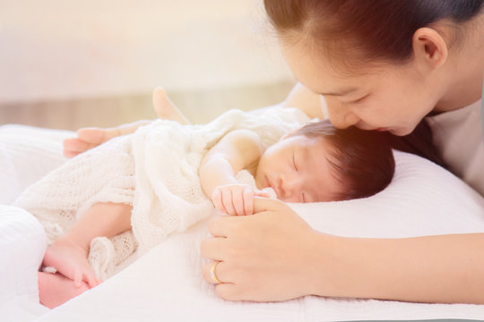 Selective Focus, Asian mother kiss adorable newborn while sleep, mom support baby girl with love, care, protection hold hand together. Motherhood lifestyle take care infant on bed at home concept