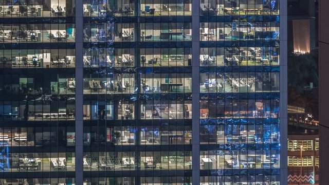 Office building exterior during late evening with interior lights on and people working inside skyscraper night timelapse. Aerial close up view from above with many illuminated windows.