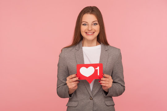 Popularity in social media. Portrait of elegant woman in business suit holding network heart Like icon and smiling to camera, recommending to follow blog. studio shot isolated on pink background