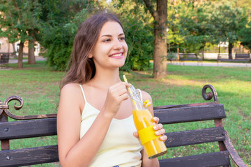 Young pretty girl is having rest on the bench in the park and drinking fresh juice from a bottle...