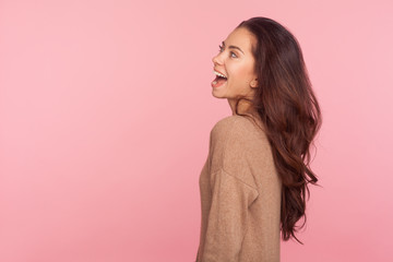 Side view of delighted slim beautiful young woman with long brunette wavy hair looking with extremely happy joyful smile, expressing optimism, positive emotions. indoor studio shot, pink background