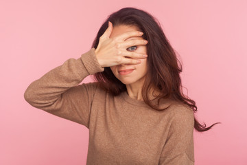 Portrait of nosy prying inquisitive young woman with brunette hair peeking through fingers with curious expression, shy and afraid to watch secret. indoor studio shot isolated on pink background