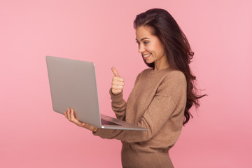 Portrait of pleased young woman in casual sweater holding laptop and showing thumbs up while talking on video call, having online conference using computer. studio shot isolated on pink background