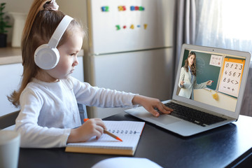 Smart small preschooler girl in headphones watch online lesson and communicate with teacher at home, little child in earphones study on Internet using laptop wireless connection.
