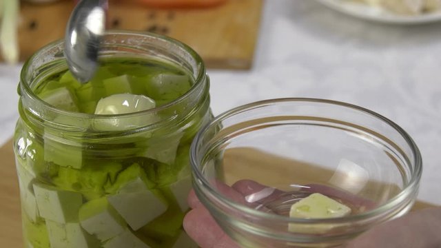 Feta cheese in olive oil is transferred from a can to a bowl with a teaspoon. Naturally transparent green olive oil contains slices of goat feta cheese.