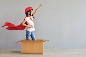 A child in a superhero costume raised his hand up while standing in a cardboard box a gray...