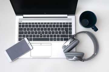 Smartphone and headphones over an open laptop on a white desk and a cup on the top right side. Top point of view