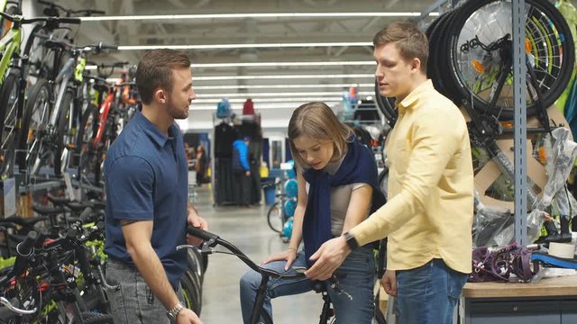 Pleasant salesman try to sell sportive bicycle for customers, smiling woman sit on sportive bicycle, listen to all characteristics.