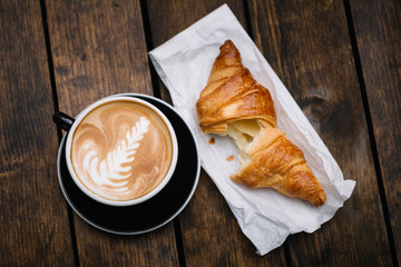 cup of cappuccino coffee with coffee art and croissant on wooden background